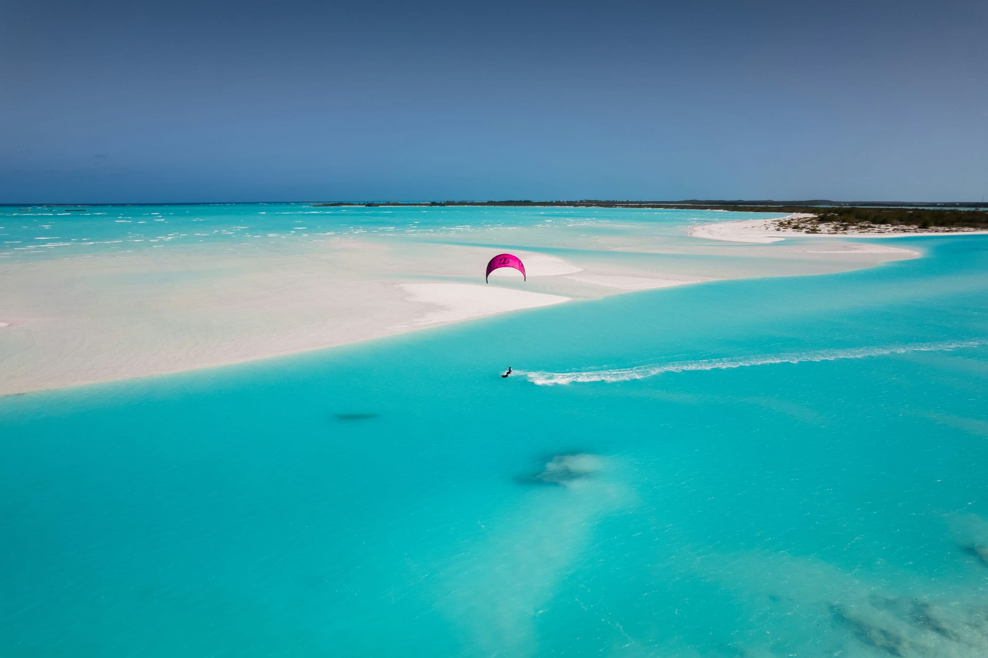 Aerial view of a lone kitesurfer under a magenta kite cutting a white wake across turquoise shallows beside pale sandbars in the Exumas, with a low green shoreline and a clear blue sky