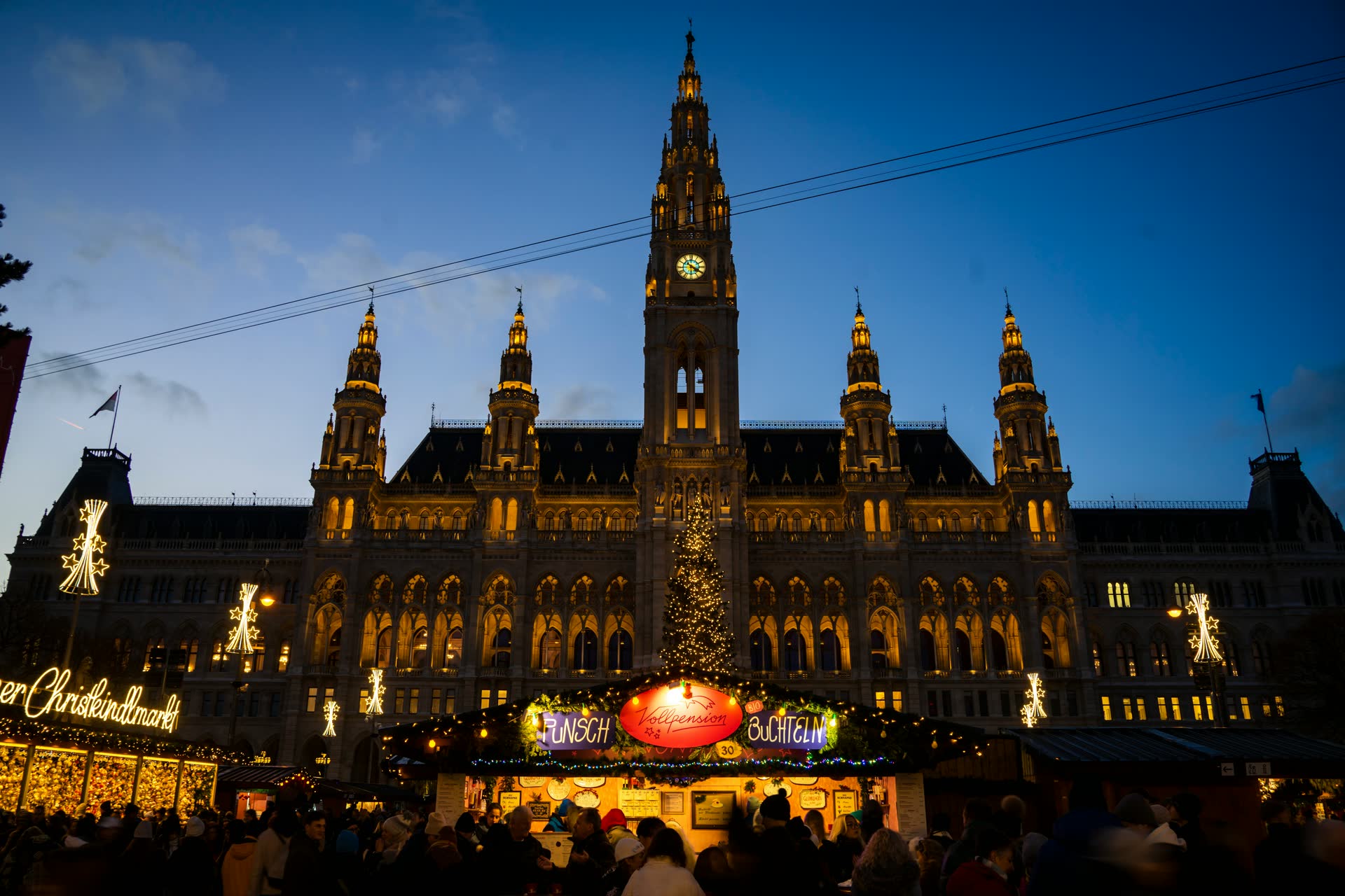 Vienna's neo-Gothic City Hall lit up at dusk behind a tall illuminated Christmas tree and a glowing market stall, with a crowd silhouetted in the foreground