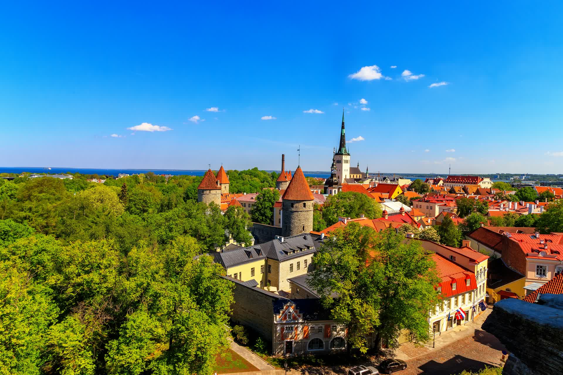 Aerial view of Tallinn's Old Town city wall towers, St. Olaf's Church spire, and red-tiled rooftops with the Baltic Sea behind