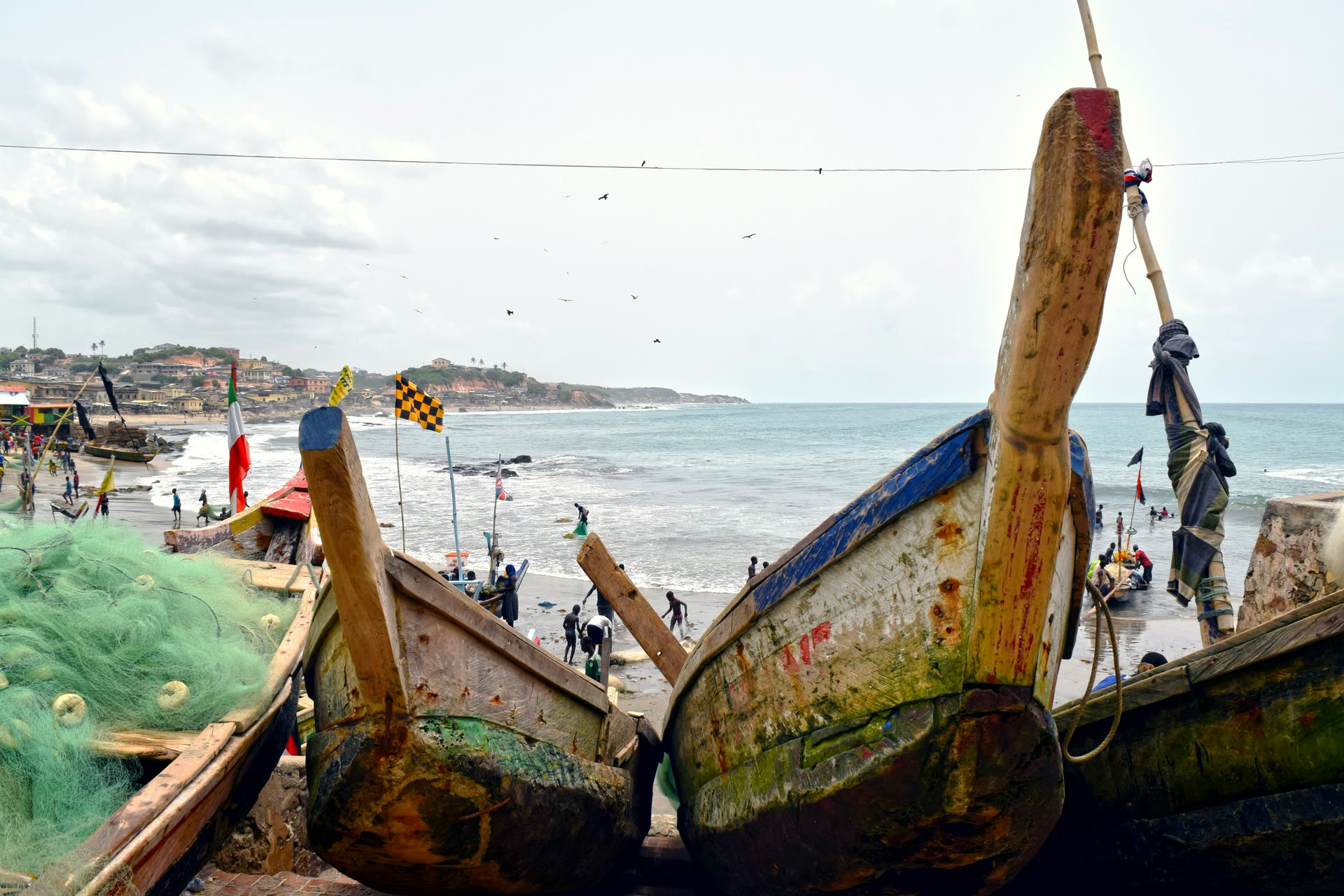 Two weathered wooden fishing canoes with blue and red trim beached side by side, green fishing nets heaped to the left, with people working at the surf line and a hillside town along the curving shore beyond, under a pale overcast sky with birds overhead