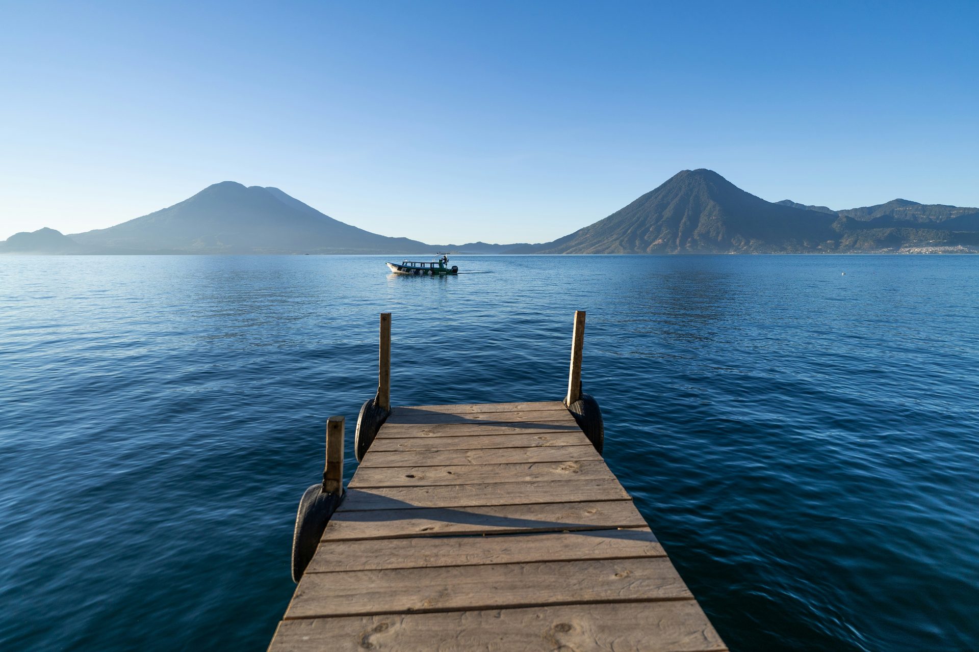 Wooden dock jutting into calm blue Lake Atitlán, with a small boat crossing the water towards two volcanic peaks under a clear sky