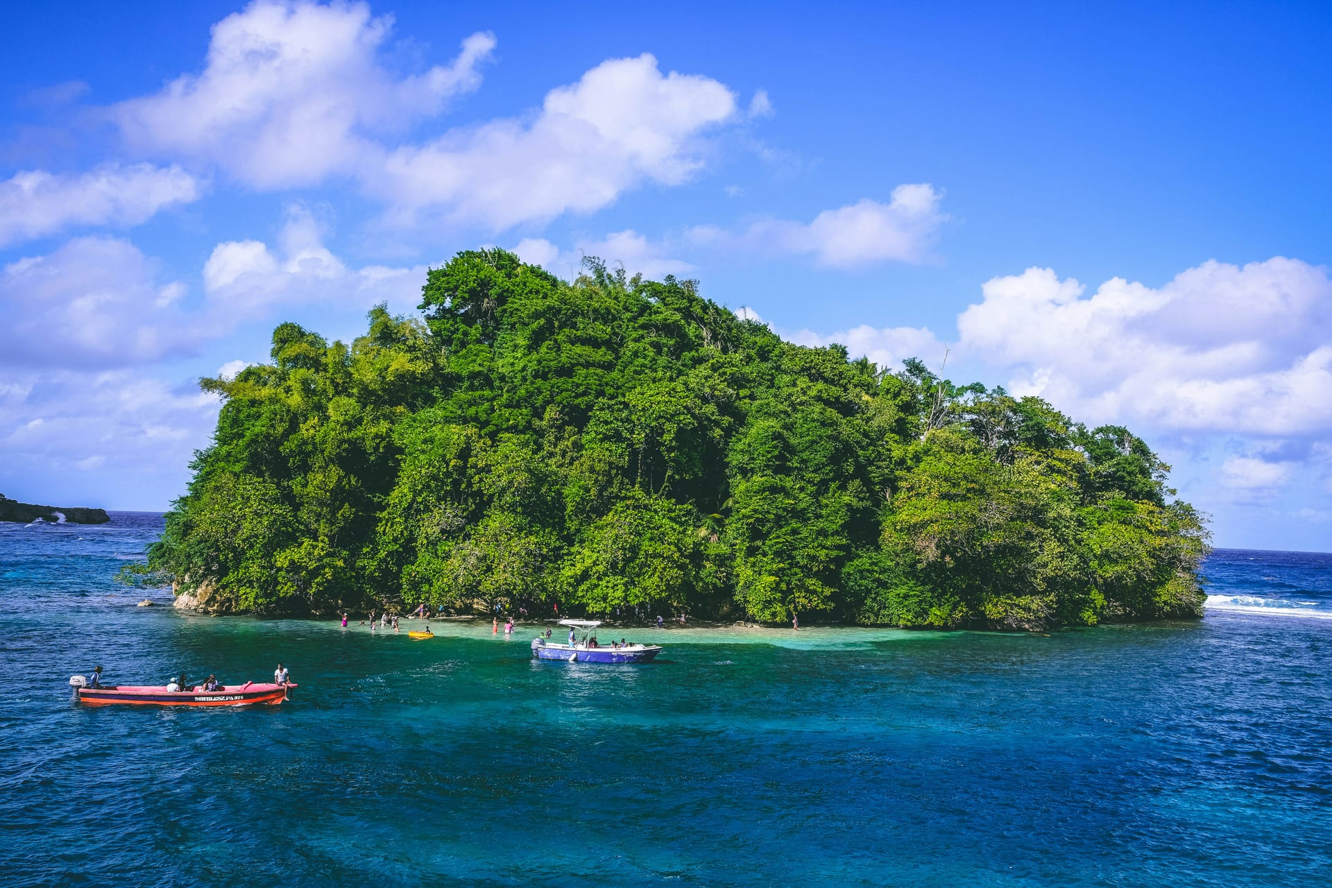 Densely wooded islet off Port Antonio, with a red boat and a blue-and-white boat on deep blue water and swimmers in the turquoise shallows, under a blue sky with white clouds