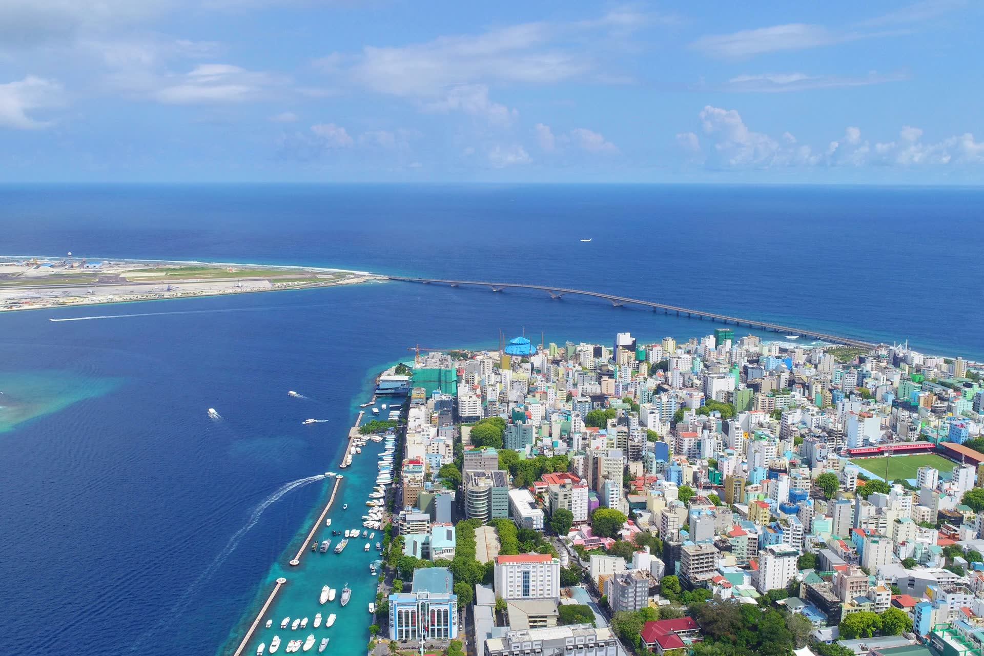 Aerial view of Malé's densely packed buildings and boat-filled harbour, with a long bridge crossing the sea to the airport island and its runway
