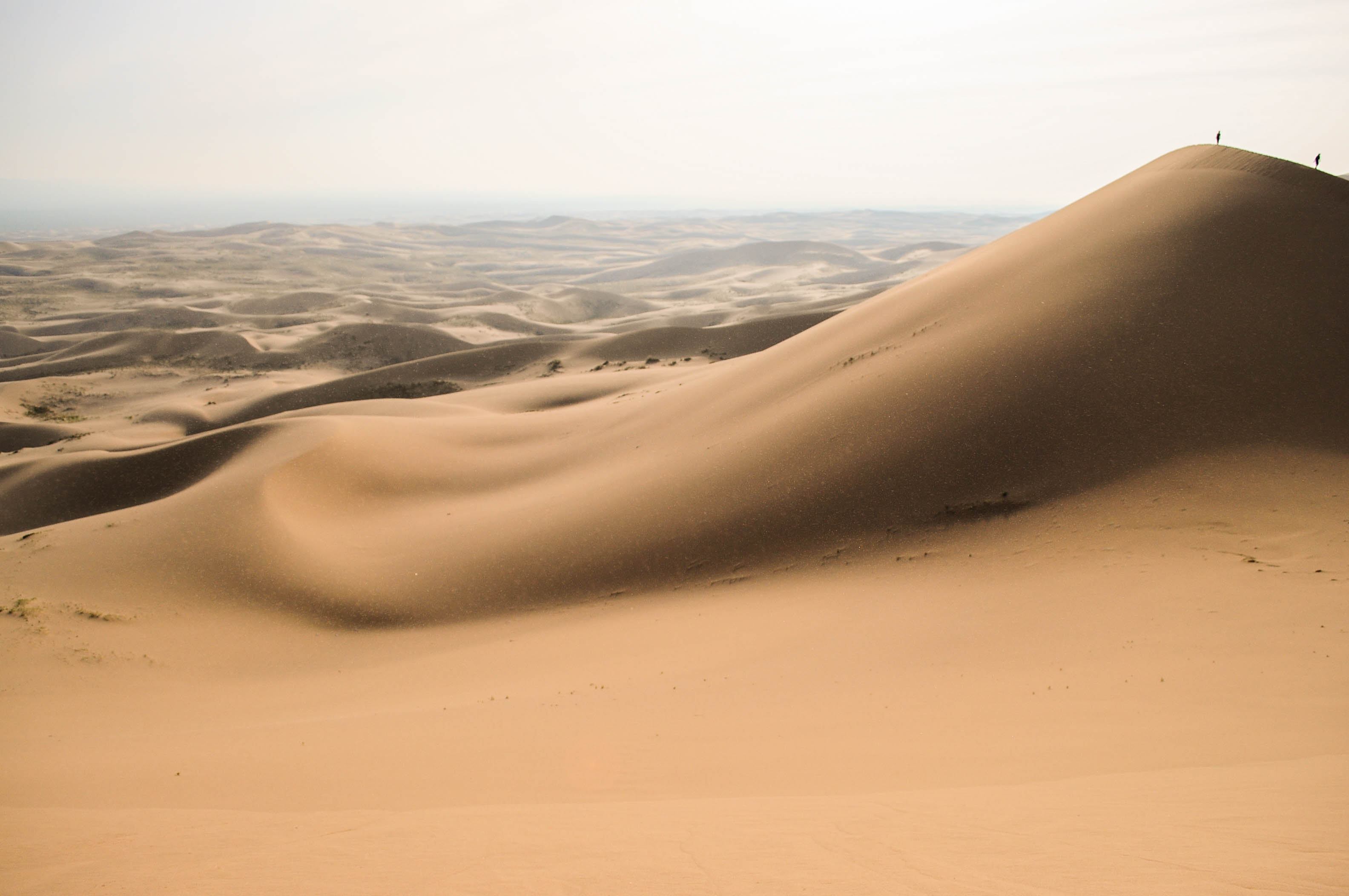 Sweeping golden sand dunes at Khongoryn Els in the Gobi Desert, with two tiny hikers on the crest of the nearest dune under a hazy sky