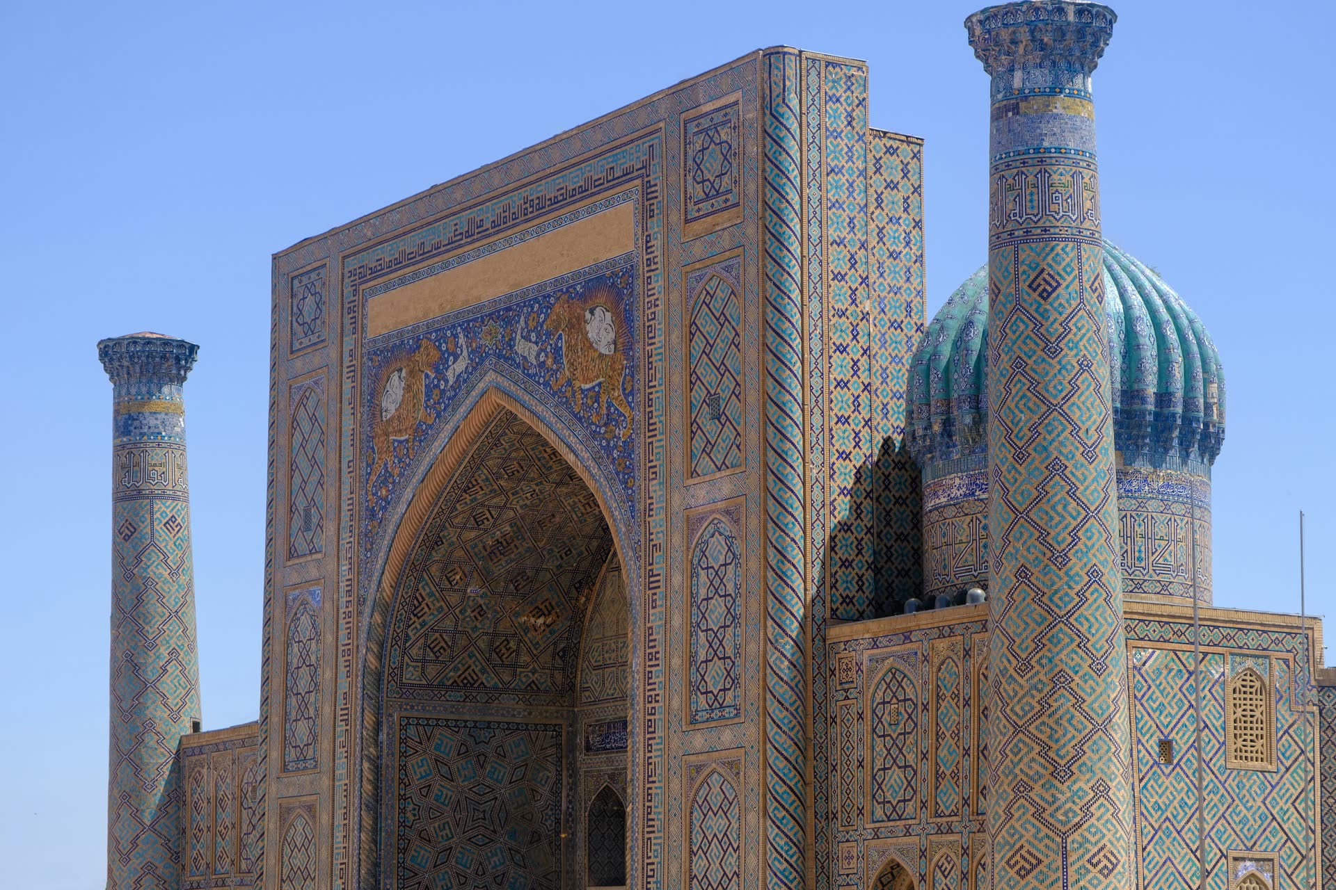 Tiled portal of the Sherdar Madrasa on Samarkand's Registan, with tiger mosaics above the arch, a ribbed turquoise dome and two minarets against a clear blue sky